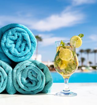 Drei aufgerollte Handtücher und ein Getränk stehen am Pool mit Palmen im Hintergrund