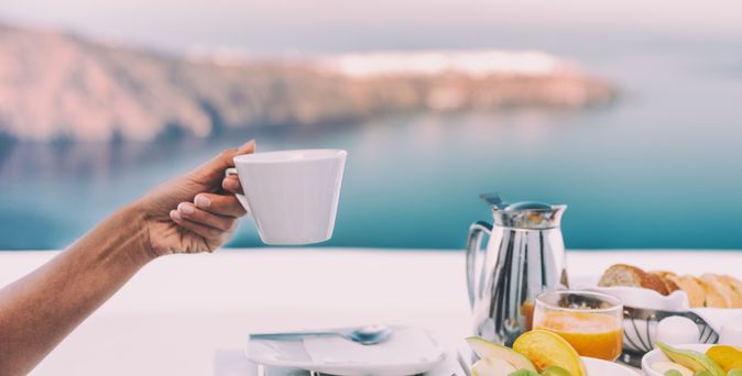 Ein gedeckter Tisch mit einer Kanne und Tellern mit Obst, eine Hand hält eine Tasse hoch, im Hintergrund ist das Meer