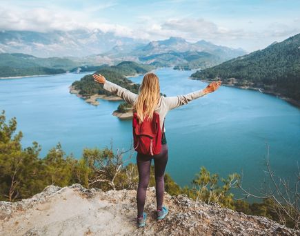 Eine Frau mit Rucksack schaut von einer Klippe auf einen großen See, eine Berglandschaft und breitet die Arme aus