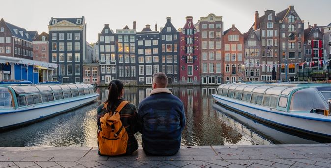 Zwei Personen sitzen in Amsterdam am Wasser und schauen gemeinsam auf die gegenüberliegenden bunten Häuser und die Boote