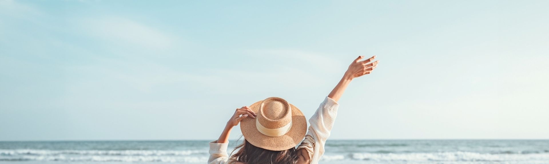 Eine Frau mit Hut von hinten, Sie schaut vom Strand aus auf das Meer und hebt den Arm 
