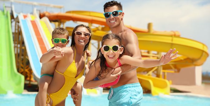 Eine Familie steht in einem Pool vor einigen Wasserrutschen, die Frau hat eins der Kinder auf dem Rücken, der Mann trägt das zweite Kind im Arm 