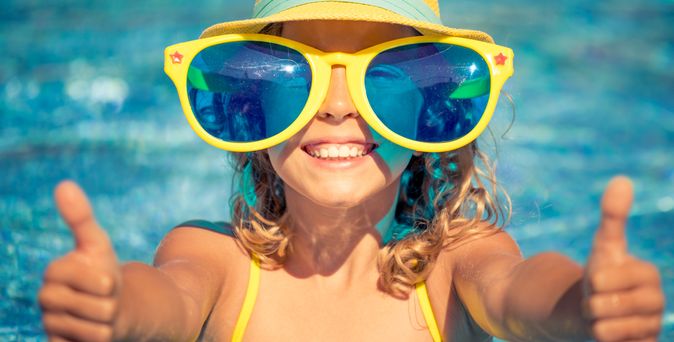 Kind mit Sonnenhut übergroßer Sonnenbrille lächelt und hält beide Daumen nach oben in die Kamera