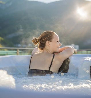 Eine Frau lehnt sich auf den Rand eines außenliegenden Whirlpools  und schaut Richtung Sonne