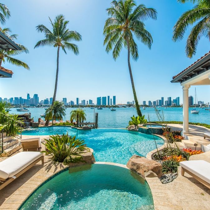 Von einem Pool umgeben von Palmen schaut man auf das Meer, im Hintergrund Hochhäuser