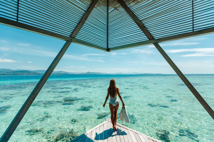 Eine Frau im Bikini steht am Ende eines Stegs und schaut aufs Meer