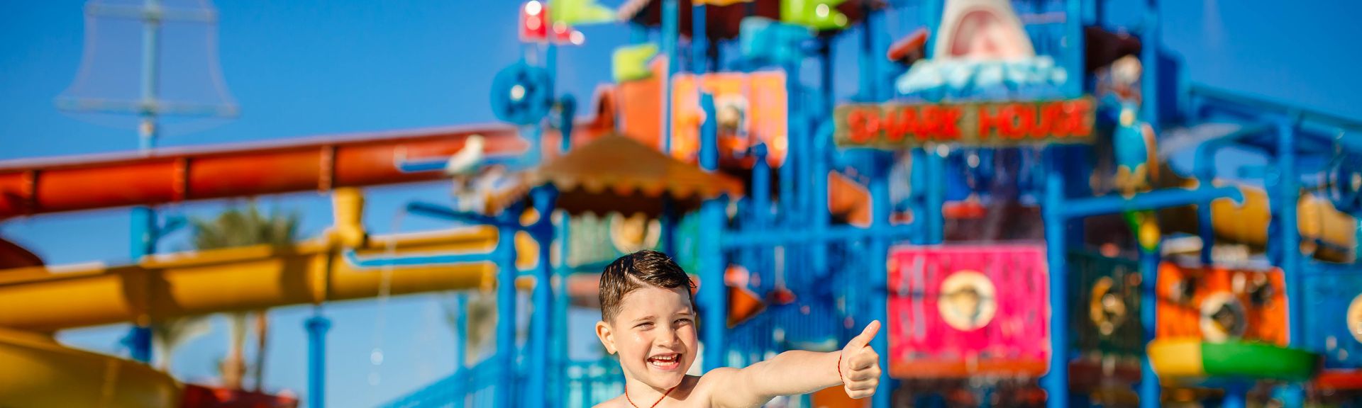 Ein Junge in einer gelben Badehosen steht vor einem großen Aquapark und zeigt einen Daumen nach oben