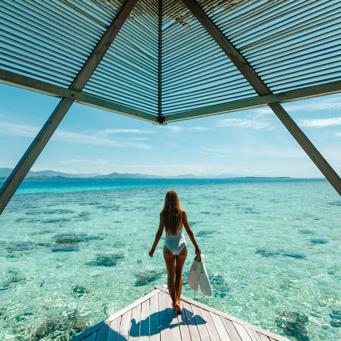 Eine Frau im Bikini steht am Ende eines Stegs und schaut aufs Meer