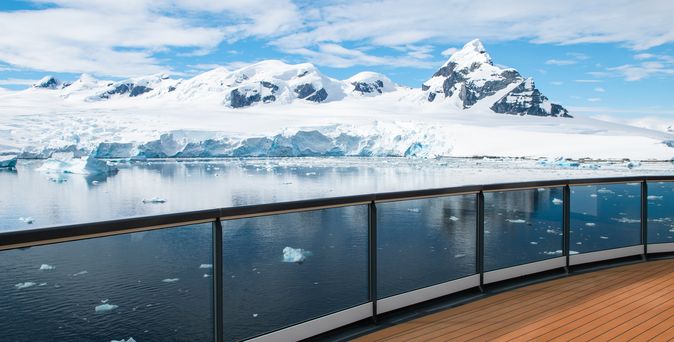 Ein Schiffsdeck mit Glasgeländer bietet einen Blick auf eine antarktische Landschaft mit schneebedeckten Bergen und schwimmenden Eisschollen