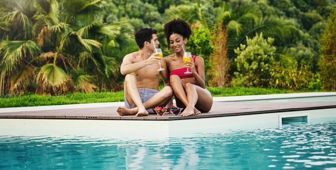 Paar sitzt mit Cocktails in der Hand und Obstschälchen auf der Terrasse vor einem Pool