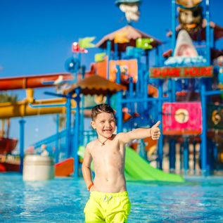 Ein Junge in einer gelben Badehosen steht vor einem großen Aquapark und zeigt einen Daumen nach oben