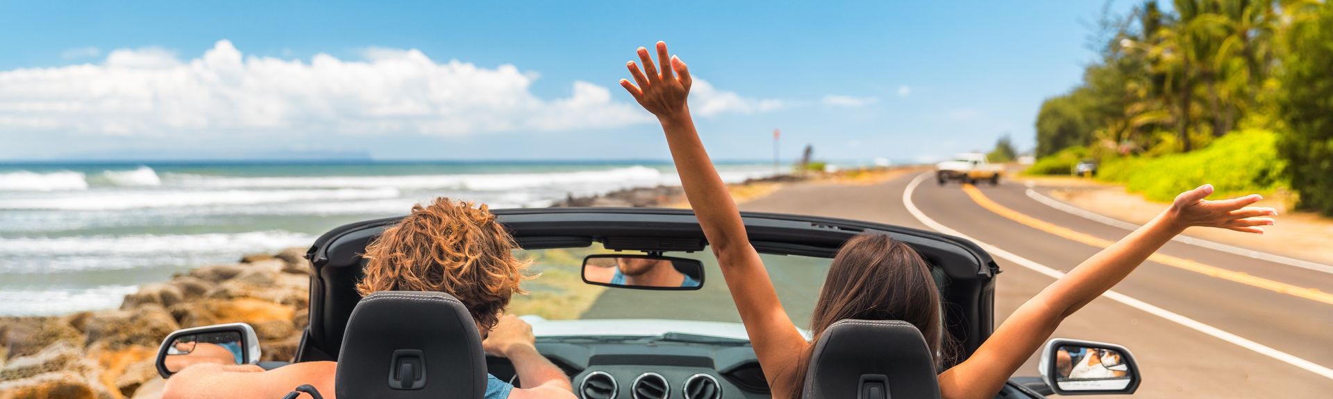 Zwei Personen im Auto mit offenem Verdeck, im Hintergrund kann man das Meer sehen