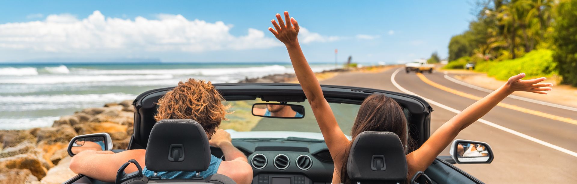 Zwei Personen im Auto mit offenem Verdeck, im Hintergrund kann man das Meer sehen