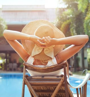 Im Hintergrund verschwommen ein Pool und ein Haus, im Vordergrund eine Frau von hinten, Sie liegt auf einer Holzliege, trägt einen Strohhut und hat die Arme hinter dem Kopf verschränkt