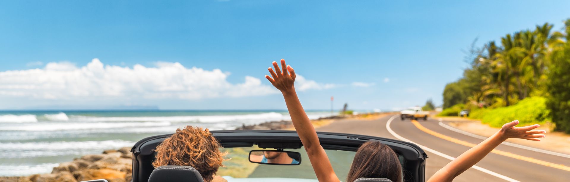 Zwei Personen im Auto mit offenem Verdeck, im Hintergrund kann man das Meer sehen