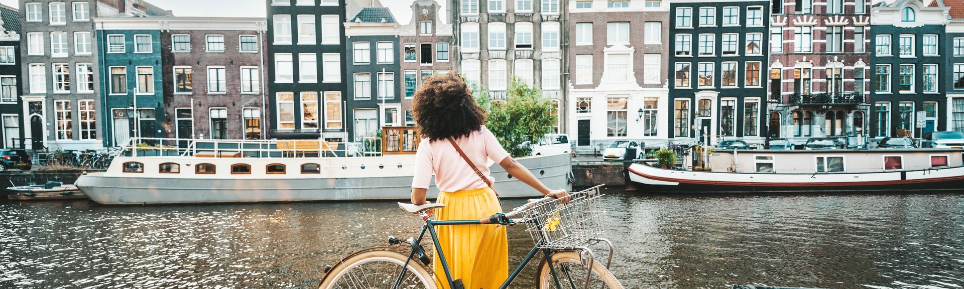 Eine Frau steht mit ihrem Fahrrad vor der Amstel und schaut auf die Häuser auf der gegenüberliegenden Seite