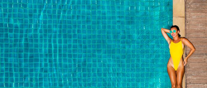 Ein großer Pool, am Rand liegt eine Frau im gelben Badeanzug und einer Sonnenbrille