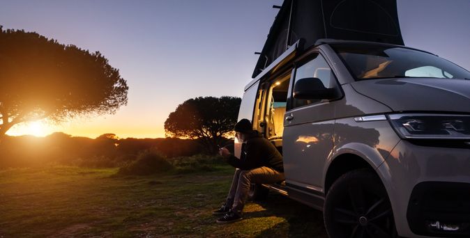 Ein Mann sitzt im Eingang eines Transporters und schaut auf sein Smartphone, im Hintergrund große Bäume und ein Sonnenuntergang