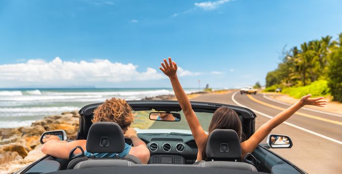 Zwei Personen im Auto mit offenem Verdeck, im Hintergrund kann man das Meer sehen