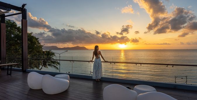 Frau am Geländer einer Terasse schaut auf den Sonnenuntergang über dem Meer