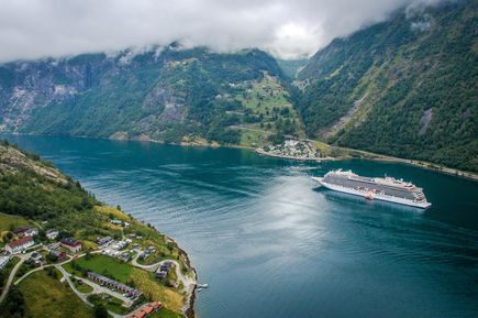 Ein Kreuzfahrtschiff fährt durch einen tiefen, von grünen Bergen gesäumten Fjord mit einem kleinen Dorf im Vordergrund.
