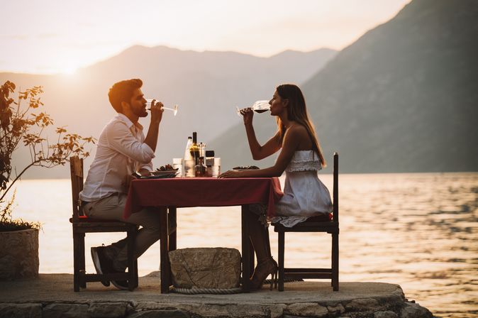 Eine Frau und ein Mann sitzen sich bei Sonnenuntergang an einem Tisch am Wasser gegenüber und trinken Wein