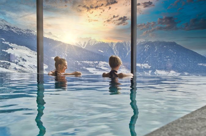 Zwei Frauen in einem Indoor Pool schauen aus dem Fenster auf schneebedeckte Berge