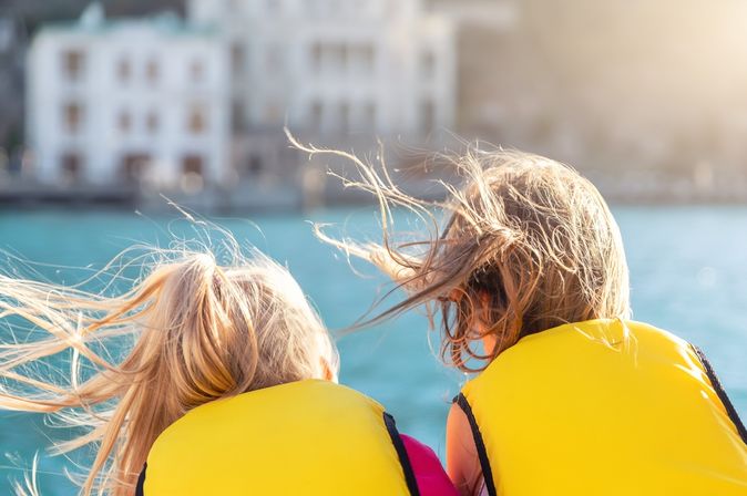 Zwei Kinder mit Schwimmwesten schauen aufs Wasser, während der Wind ihre Haare verweht