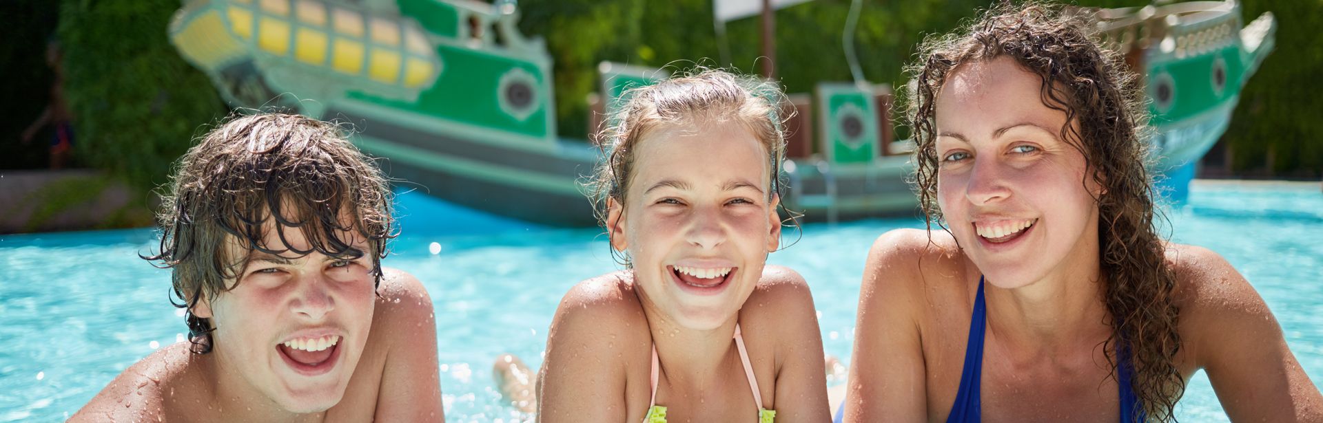 Zwei Mädchen und ein Junge lehnen am Rand eines Pools und lächeln in die Kamera, im Hintergrund sieht man ein Piratenschiff