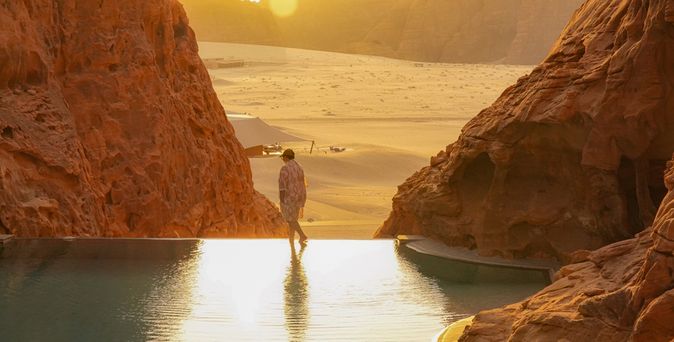 Person läuft am Rand eines Infinity Pools entlang, der zwischen Felsen steht. Im Hintergrund ist Sand und ein Sonnenuntergang zu sehen