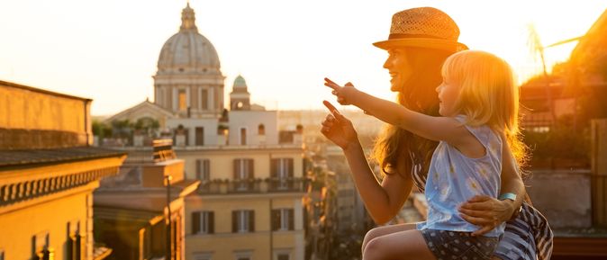 Eine Frau sitzt auf einer Mauer mit Ausblick auf eine Stadt. Auf ihrem Schoß sitzt ein kleines Mädchen und sie zeigen gemeinsam auf ein Bauwerk 