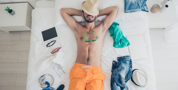 Ein Mann in oranger Badehose liegt auf dem Rücken im Bett, sein Hut liegt auf seinem Gesicht und um ihn herum liegen Anziehsachen, ein Tablet und eine Sonnenbrille