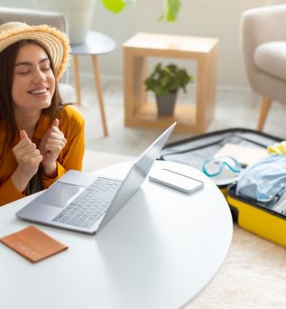 Eine Frau sitzt glücklich vor ihrem Laptop, auf dem Tisch liegen Reisepass und Kreditkarte, im Hintergrund liegt ein gepackter Koffer