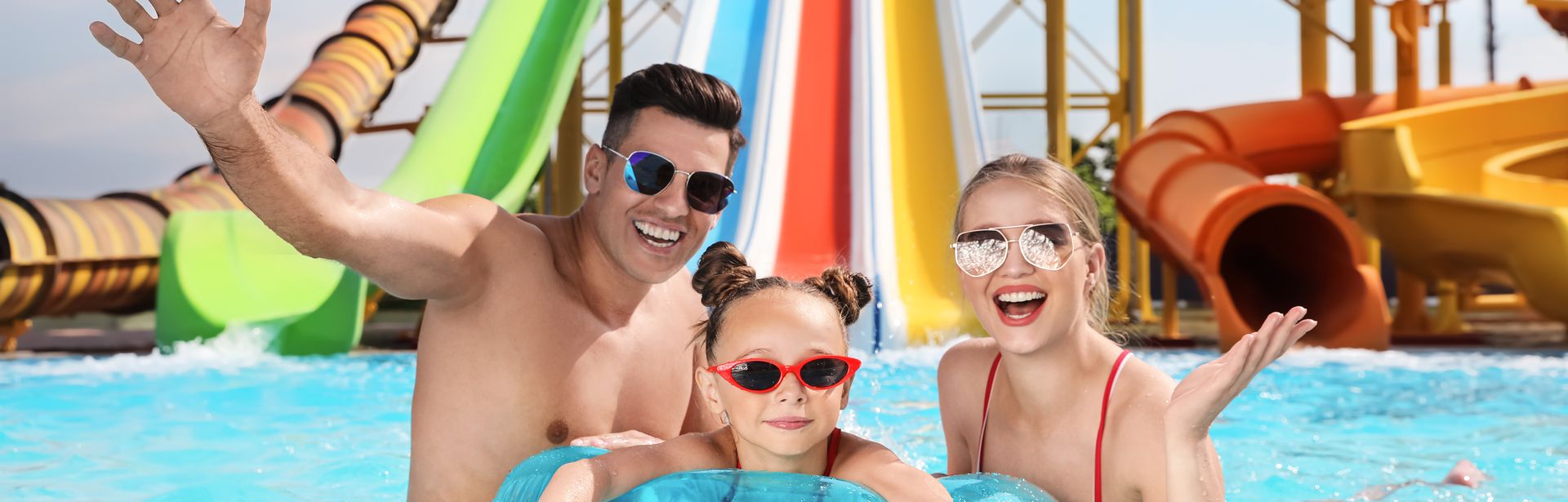 Eltern und ein Kind in einem Pool, Das Kind trägt eine Sonnenbrille und schwimmt in einem Schwimmring, im Hintergrund sind Wasserrutschen