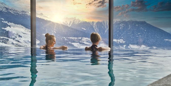 Zwei Frauen in einem Indoor Pool schauen aus dem Fenster auf schneebedeckte Berge