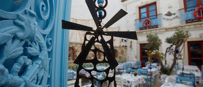 Ein Windspiel in Form einer Windmühle hängt in einem Hinterhof mit bunt gedeckten Tischen und blauen und weißen Stühlen