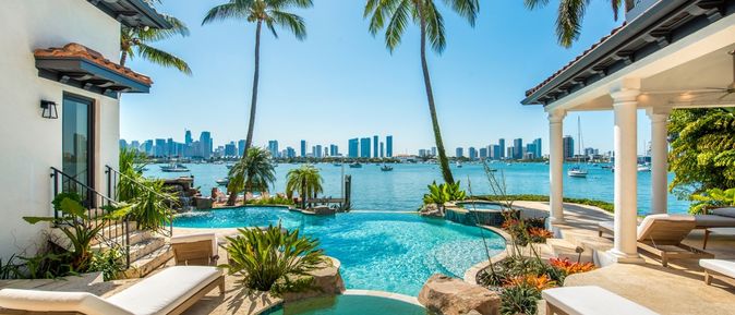 Von einem Pool umgeben von Palmen schaut man auf das Meer, im Hintergrund Hochhäuser