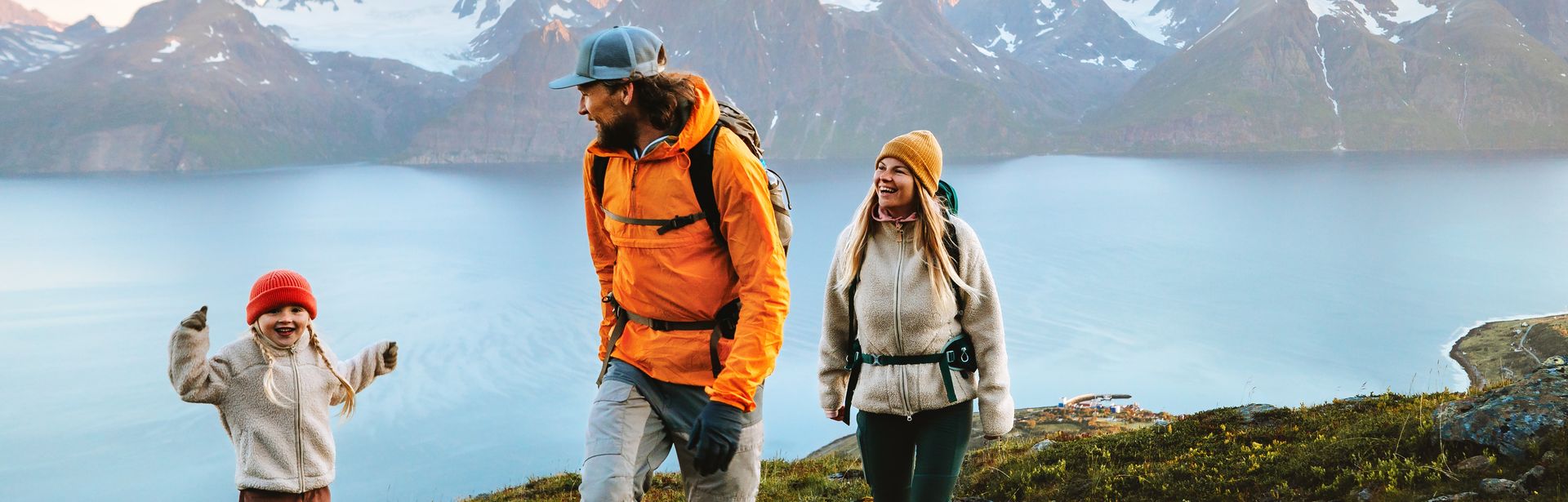 Eine Familie wandert in Winterkleidung an einem Gewässer, im Hintergrund sind verschwommen Berge 
