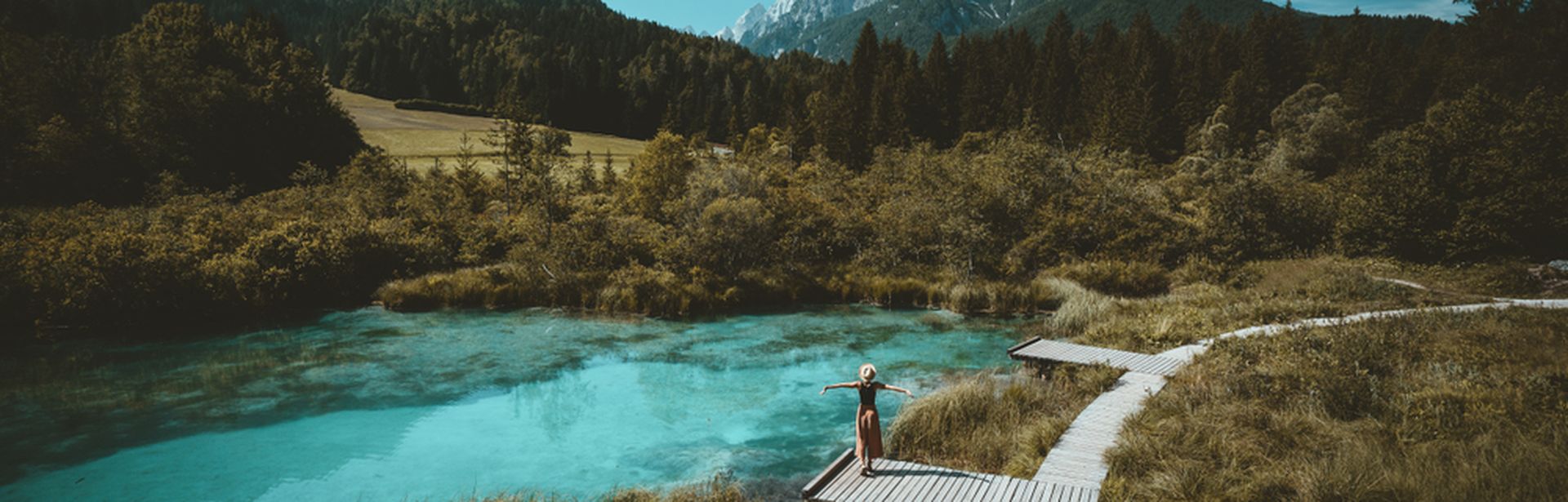 Eine Große Wiese mit einem Holzweg, auf einem Steg steht eine Frau und breitet die Arme aus, sie schaut auf den See vor ihr