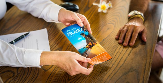 Ein Schreibtisch in einem Reisebüro, der Reiseberater überreicht eine Broschüre zu einem Urlaubsland
