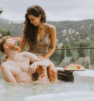 Ein Mann sitzt im Whirlpool und schaut lachen zu einer Frau, die am Rand des Whirlpools über ihm sitzt