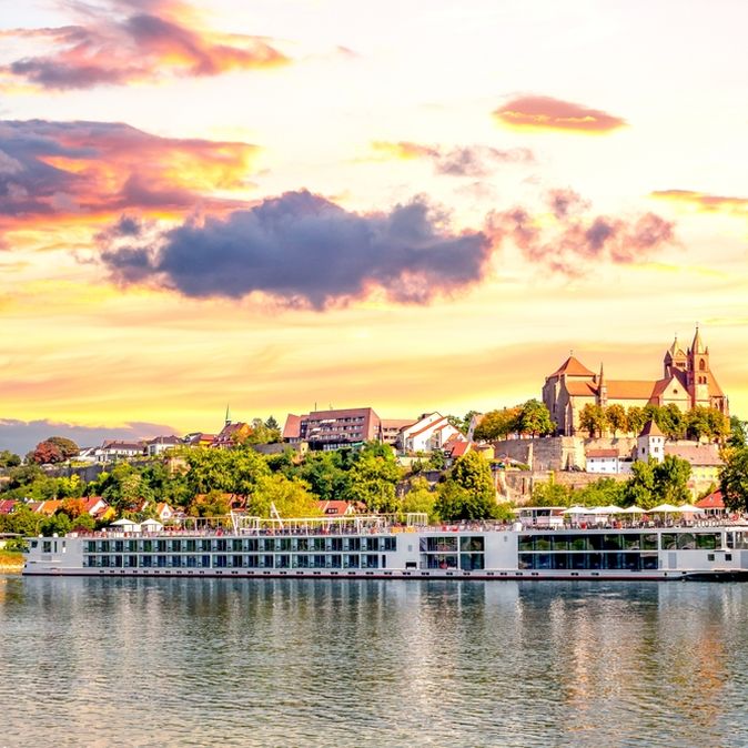 Langes Flusskreuzfahrschiff fährt entlang eines kleinen Dorfes, bei gelb leuchtendem Himmel