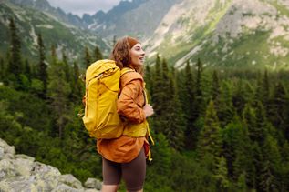 Eine Frau mit gelbem Wanderrucksack steht auf einem Berg und schaut begeistert in die Ferne