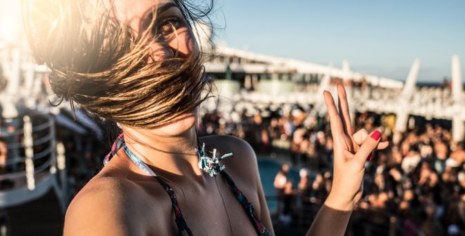Eine Frau macht auf einem Kreuzfahrtschiff ein Selfie, Ihre Haare wehen im Wind und im Hintergrund sieht man eine feiernde Menge