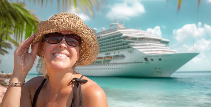 Eine Frau mit einem Strohhut lacht in die Kamera, im Hintergrund sieht man ein Kreuzfahrtschiff