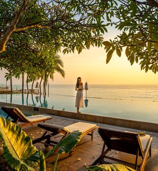 Frau steht bei Sonnenuntergang vor einem Pool der direkt vor einem Meer liegt