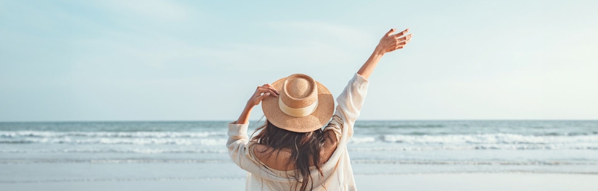 Eine Frau mit Hut von hinten, Sie schaut vom Strand aus auf das Meer und hebt den Arm 