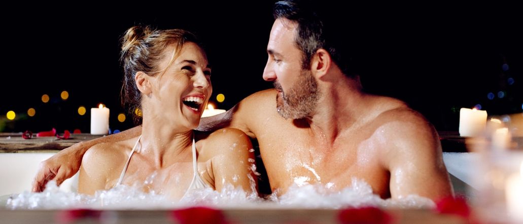 Ein Paar sitzt lachend in einem Whirlpool bei Kerzen und Rosenblättern