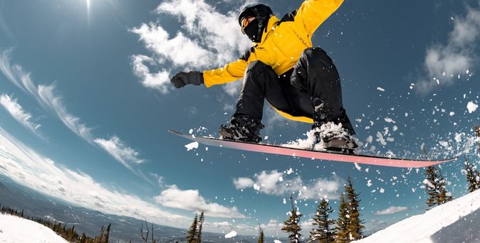 Eine Person in gelber Winterjacke springt mit einem Snowboard durch die Luft