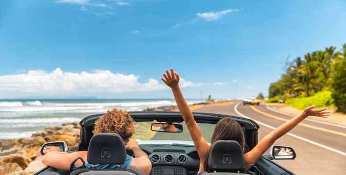 Zwei Personen im Auto mit offenem Verdeck, im Hintergrund kann man das Meer sehen